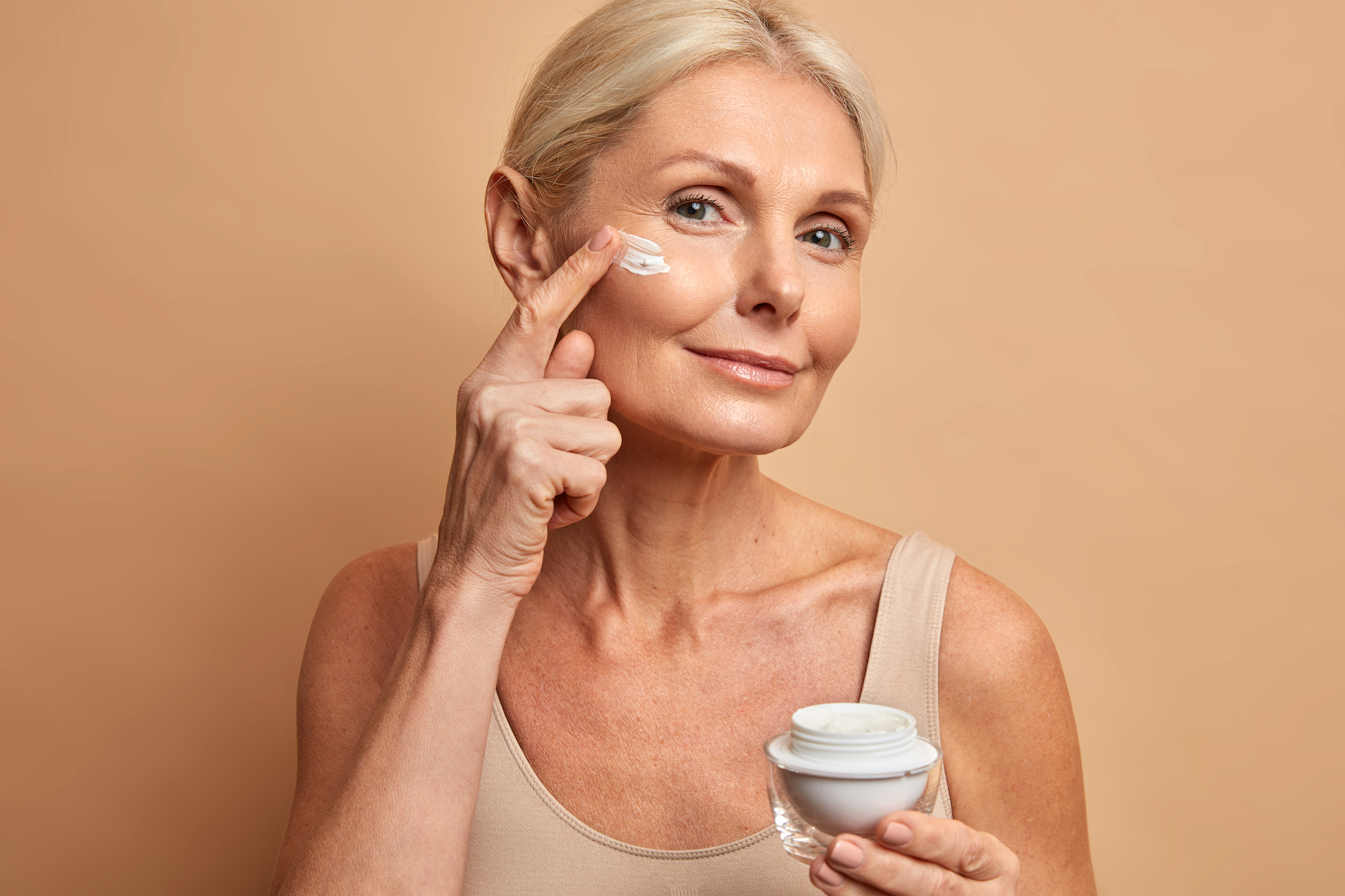 Close up shot of middle aged beautiful woman applies anti aging cream on face undergoes beauty treatments cares about skin poses against beige background. Wrinkled female model with cosmetic product